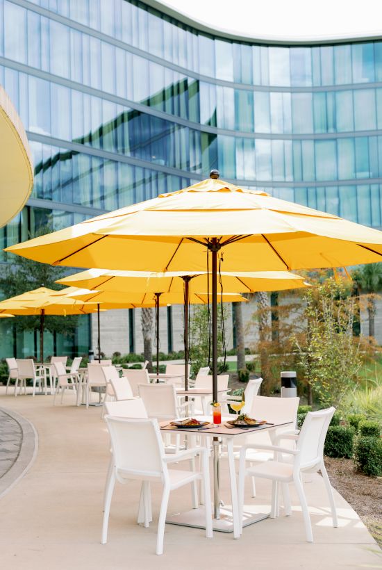 A modern outdoor patio with white tables and chairs under large yellow umbrellas, beside a glass-front building.