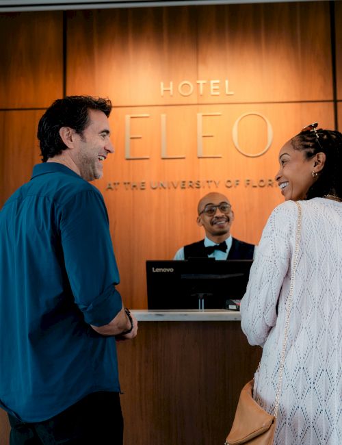 Two guests chat with a receptionist at a hotel front desk; warm wood backdrop reads "HOTEL LEO" with flowers on the side.