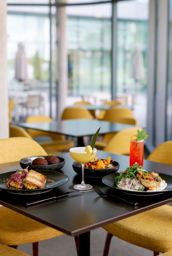 A stylish dining setup with four plated dishes and colorful cocktails on a black table in a bright, modern restaurant with yellow chairs and large windows.