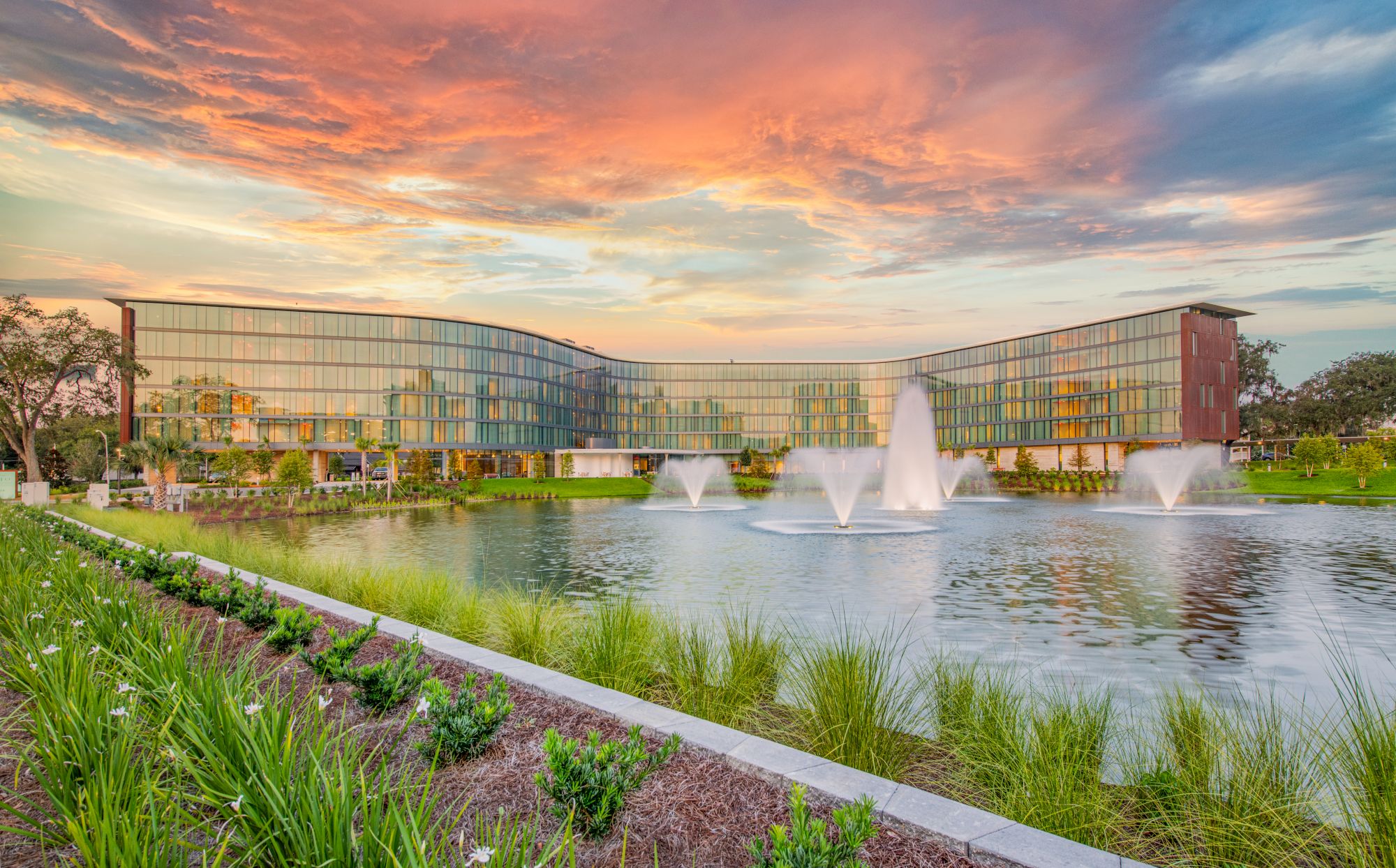 A modern campus with glass-front buildings around a central lake, trimmed by fountains and landscaped gardens at sunset.