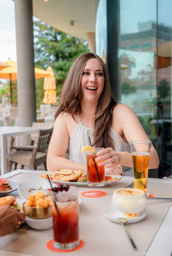 Three friends chat and laugh over drinks on an outdoor patio, plates of food and colorful beverages on the table.