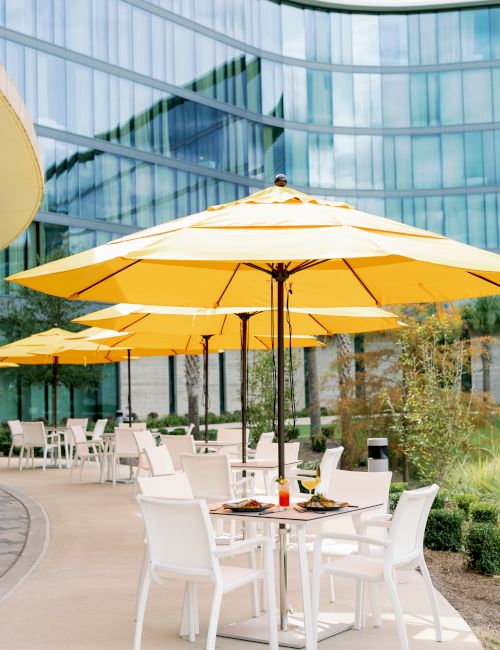 Outdoor cafe setting with white tables and chairs under yellow umbrellas, modern glass building backdrop, daylight, relaxed dining vibe.