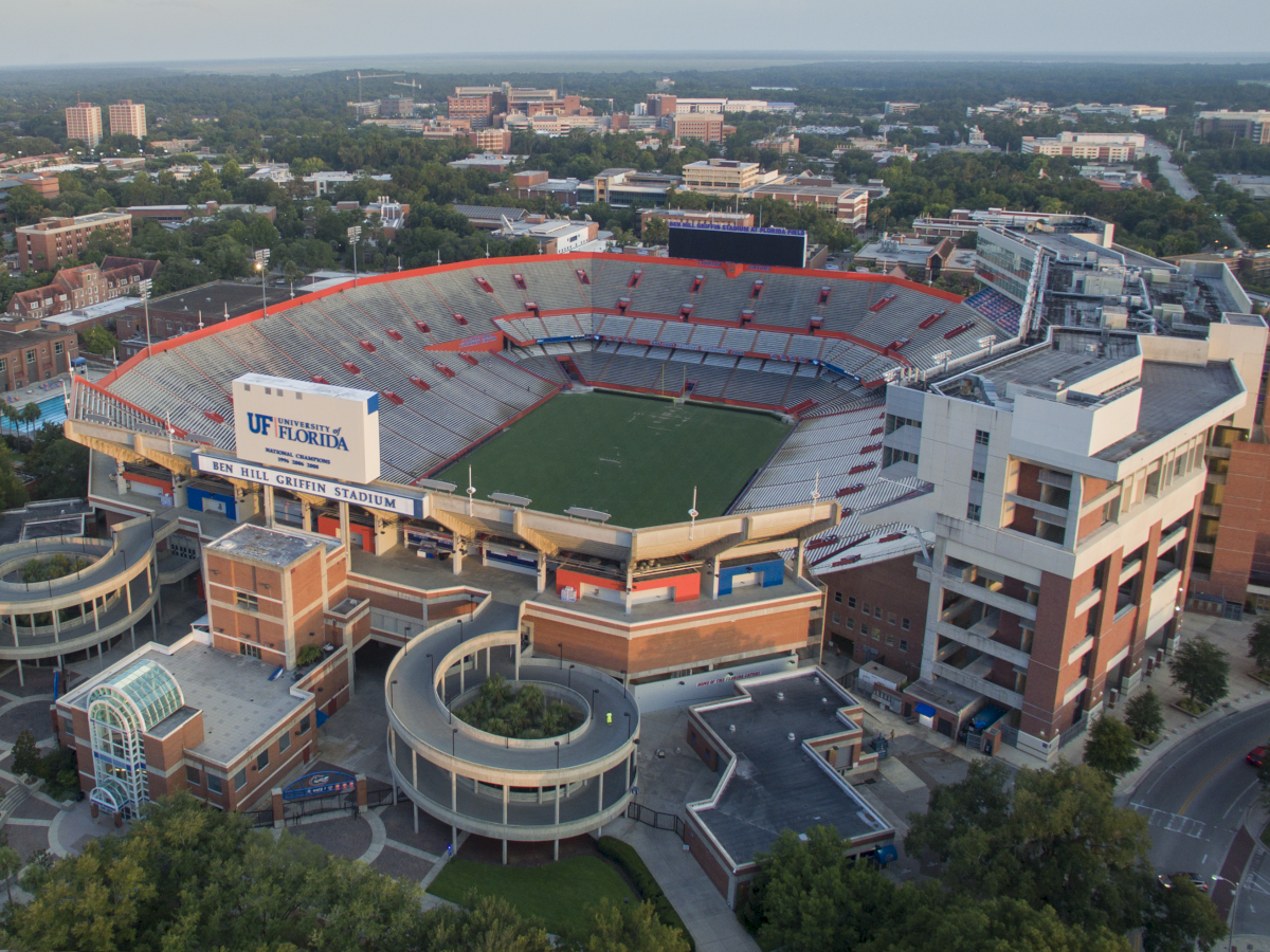 An aerial view of a large college football stadium with surrounding buildings, greenery, and a rectangular field ready for a game, under clear skies.