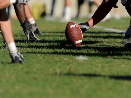 A football rests on a grassy field as players in gear prepare for a game and a kickoff, with legs and hands visible everywhere.
