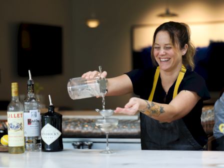 A bartender smiles while pouring a drink into a martini glass, with liquor bottles and a bar setup in the foreground.