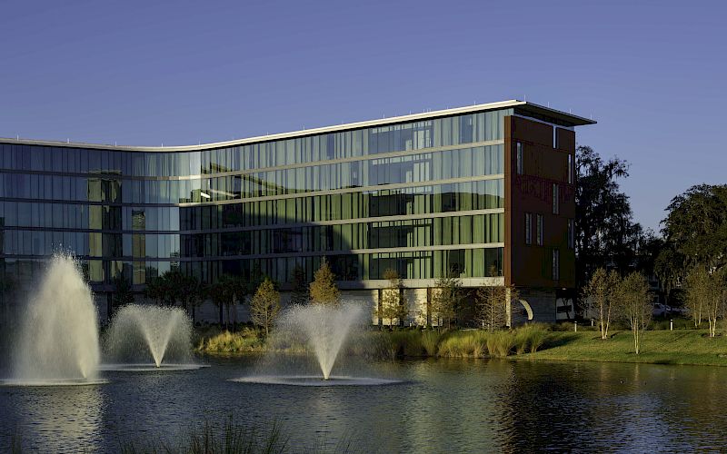 Modern glass office building with a red tower, reflected by a pond with fountains under a clear blue sky.