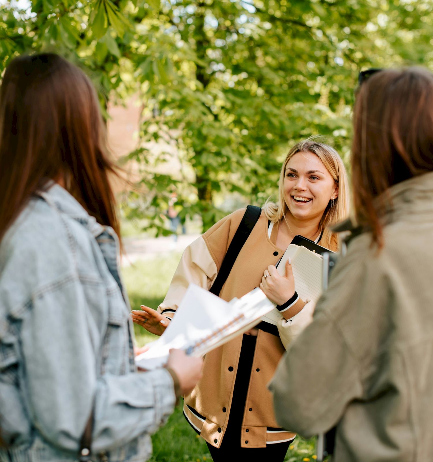 A smiling blonde woman chats with two friends in a sunny park, holding notebooks and papers, surrounded by lush green trees.