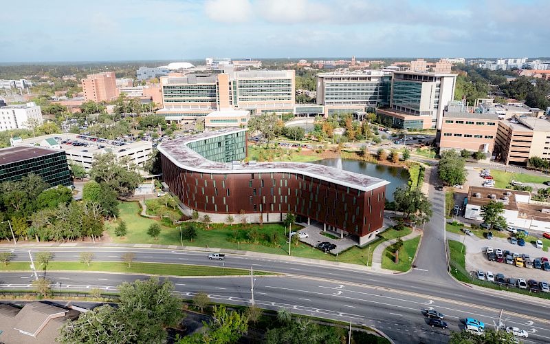 Aerial view of a modern university campus with red-brick buildings, offices, trees, parked cars, and a highway outside.