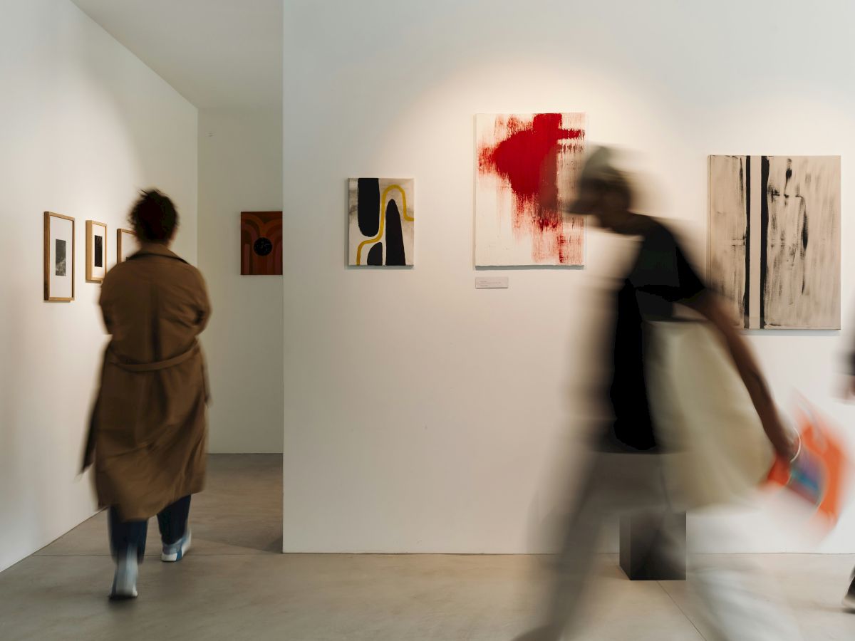 A gallery wall of colorful abstract paintings with blurred visitors walking past, capturing a lively art-viewing moment.