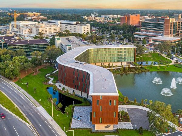 Aerial view of a modern brick concert hall beside a pond with fountains, roads, and campus buildings in the background.