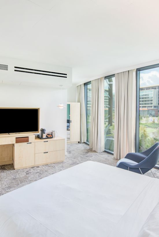 Modern hotel bedroom with large windows, curtains, bed, and TV on a light wood console, overlooking city.
