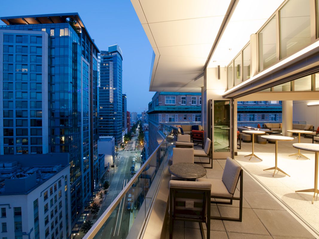 The image shows a modern cityscape with tall buildings and a sophisticated rooftop patio featuring tables, chairs, and an open indoor sitting area.