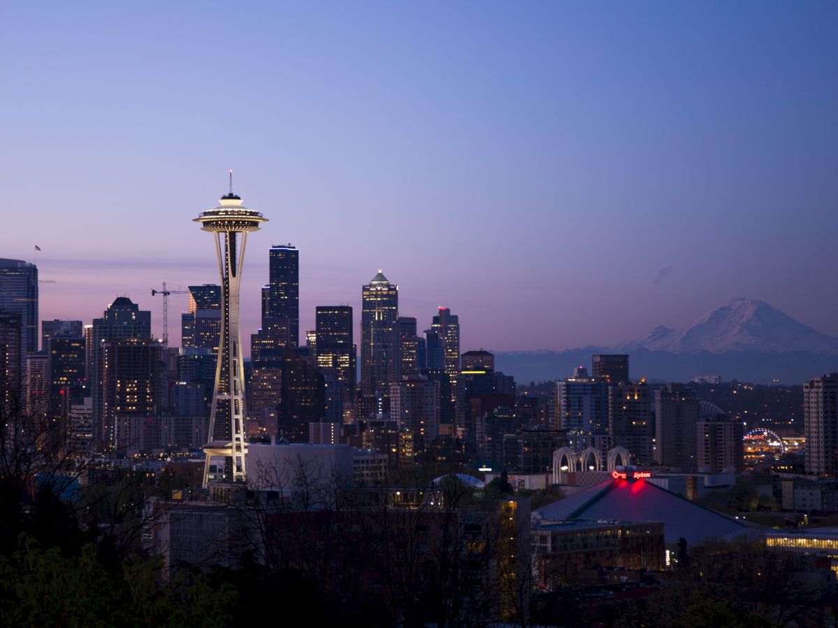 This image shows a cityscape at dusk, with a prominent tower in the foreground and a mountain in the background, under a clear evening sky.