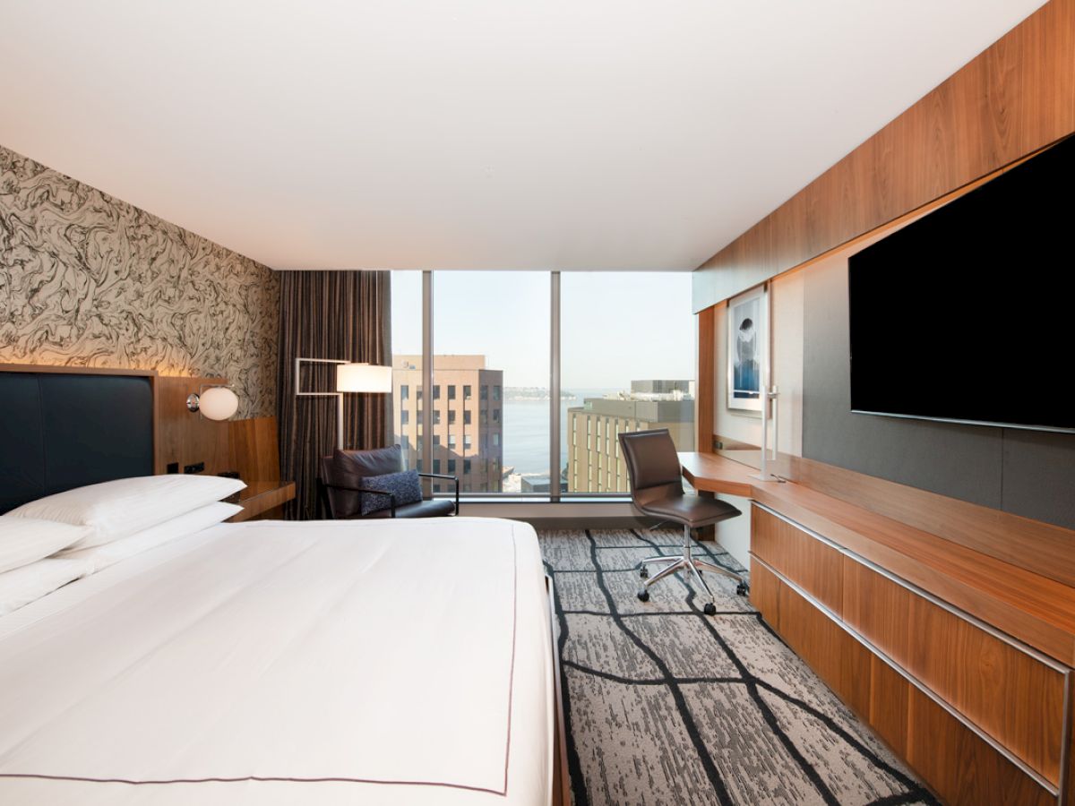 A modern hotel room with a large bed, a flat-screen TV, a desk, and a city view through large windows is shown.