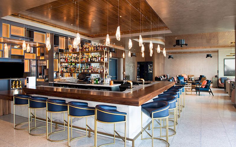 A modern bar with blue chairs, hanging lights, and a well-stocked shelf of liquor bottles in a spacious, stylish setting.